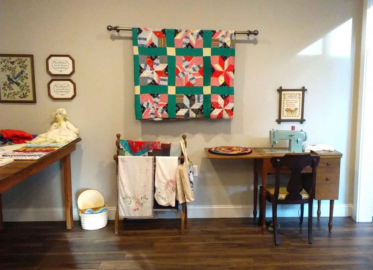 A cozy craft room with a vibrant quilt wall hanging, vintage furniture, and sewing materials.