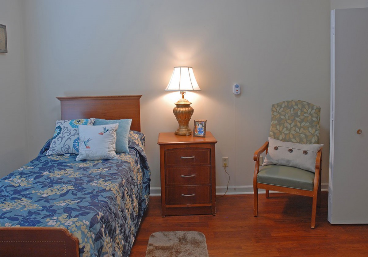 A cozy bedroom with a bed, nightstand, and chair, softly lit by a table lamp.