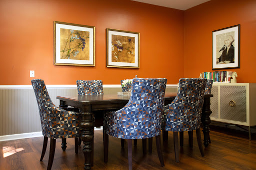 A vibrant dining room featuring a wooden table surrounded by patterned chairs and adorned with framed artwork on an orange wall.