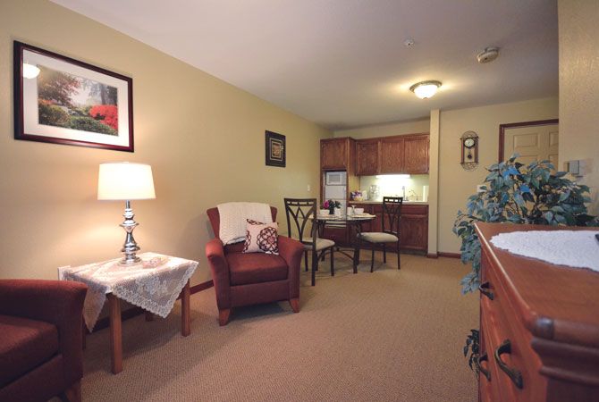 A cozy apartment space featuring a small living area with a chair, lamp, and table, leading into a compact kitchen.