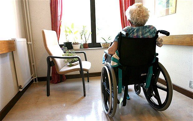 An elderly person in a wheelchair looking out of a window in a nursing home.