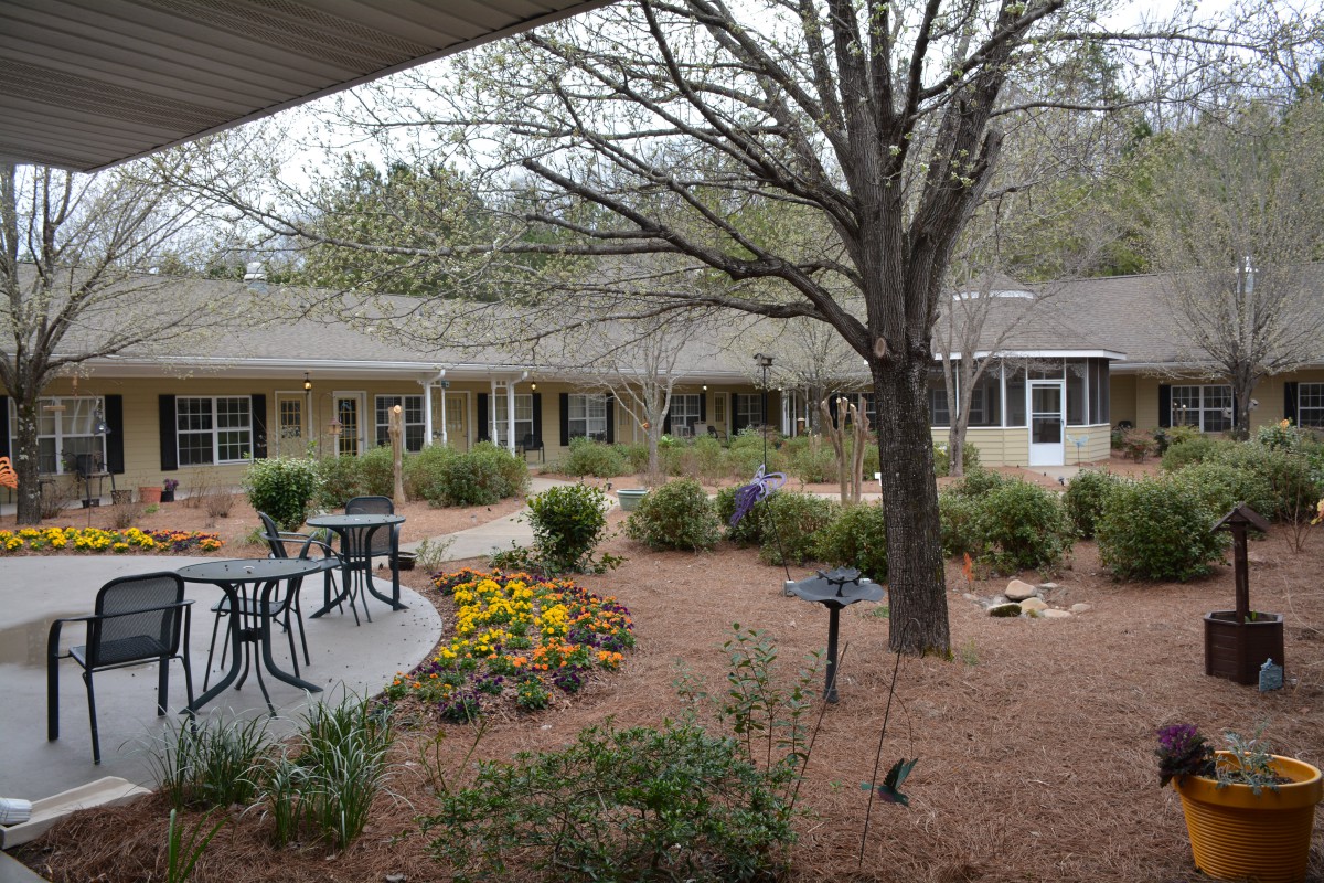 A peaceful garden with a patio featuring tables and chairs, surrounded by trees and landscaping.
