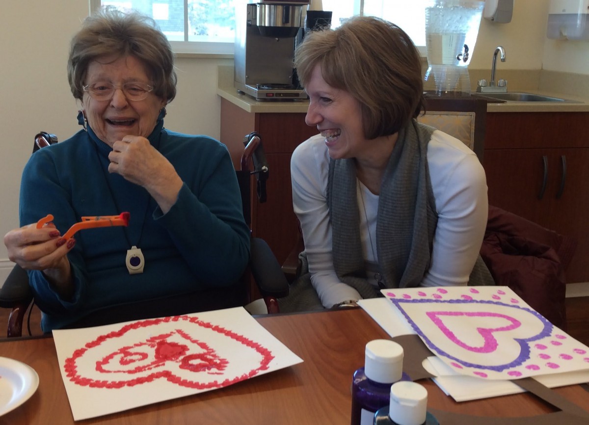 An elderly woman and a younger woman share a laugh while doing art projects at a table indoors.