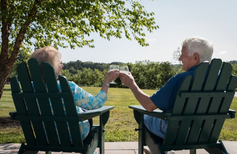 An elderly couple enjoying a peaceful moment outdoors, toasting with drinks while seated in chairs overlooking a scenic landscape.