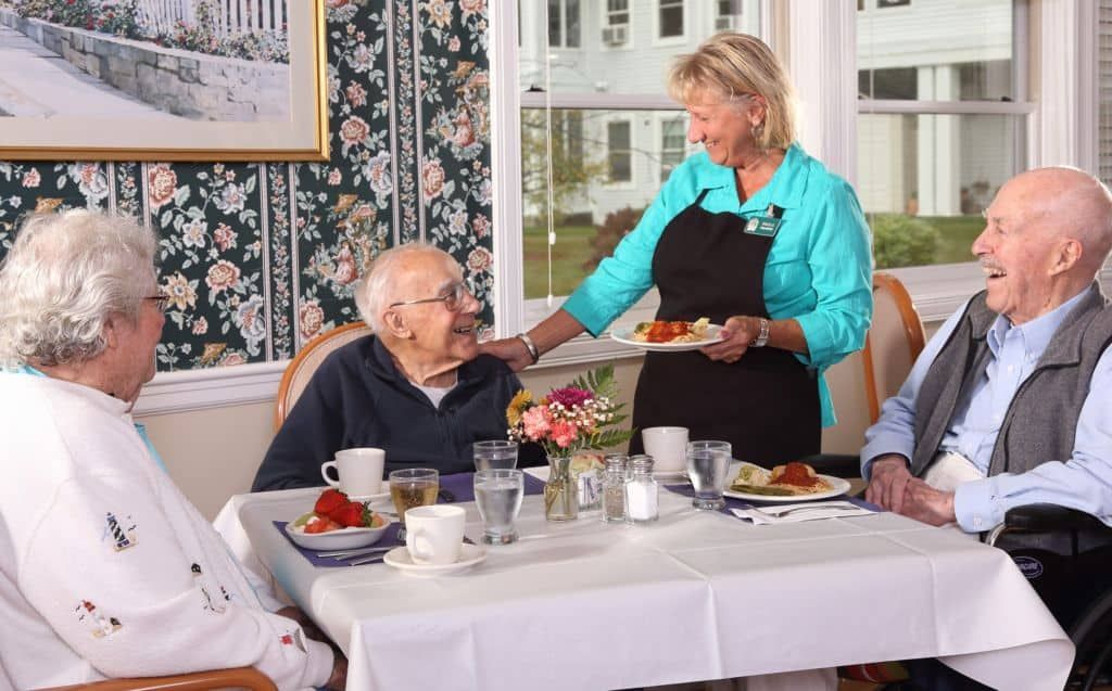 A caregiver serves a meal to an elderly person at a dining table in a warm, welcoming environment.