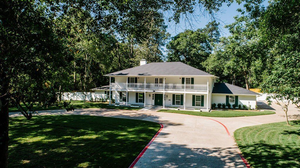 A two-story residential house with a spacious driveway surrounded by trees and greenery.