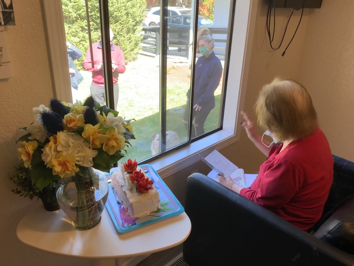 An elderly woman celebrates her birthday indoors with a cake and flowers, while family or friends visit from outside the window.