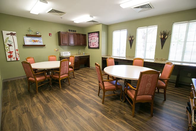 A cozy dining room with round tables and cushioned chairs, featuring wooden flooring and ample natural light from large windows.