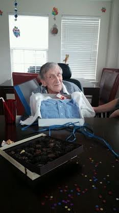 An elderly person sitting in a wheelchair at a table with an open box of chocolates nearby.