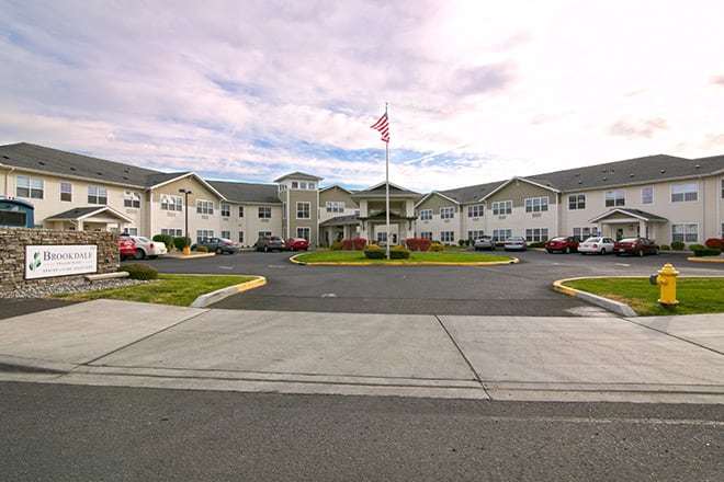 A residential building with a flagpole in front, surrounded by a parking lot and landscaping under a cloudy sky.