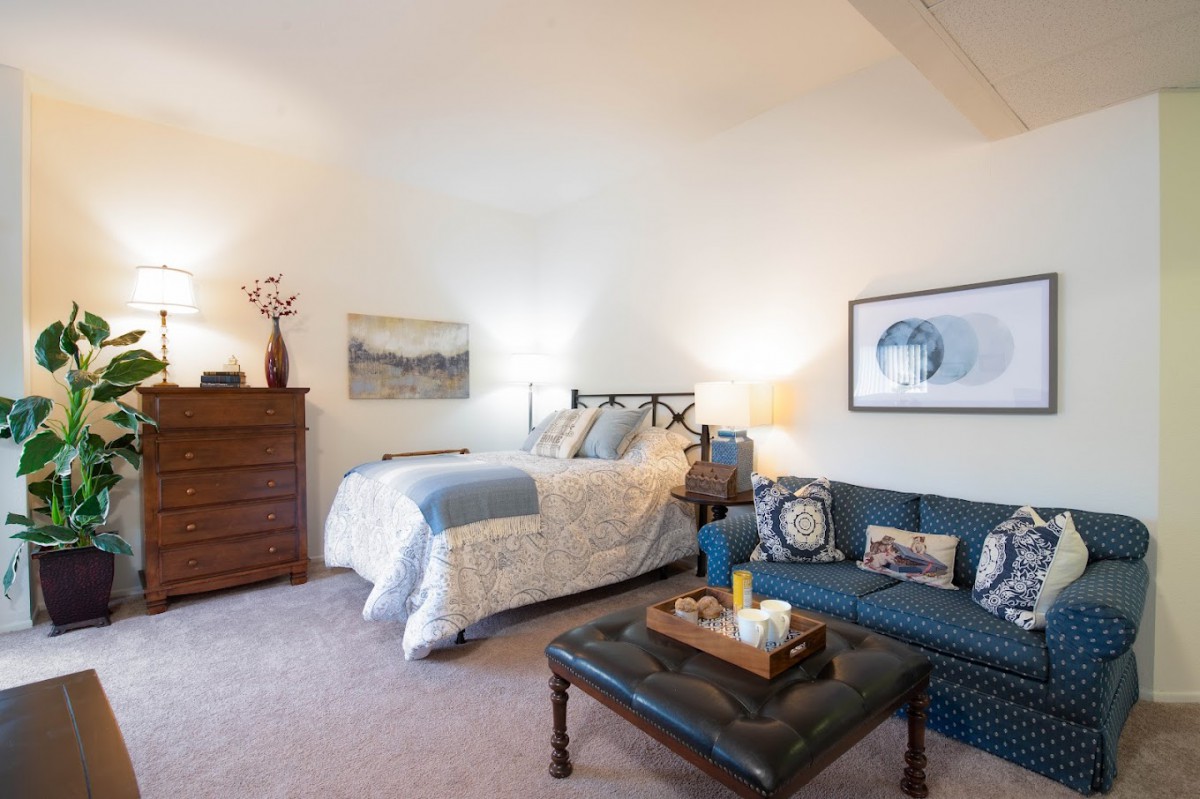 A cozy bedroom with a bed, a blue couch, and decorative elements, including artwork and a wooden dresser.