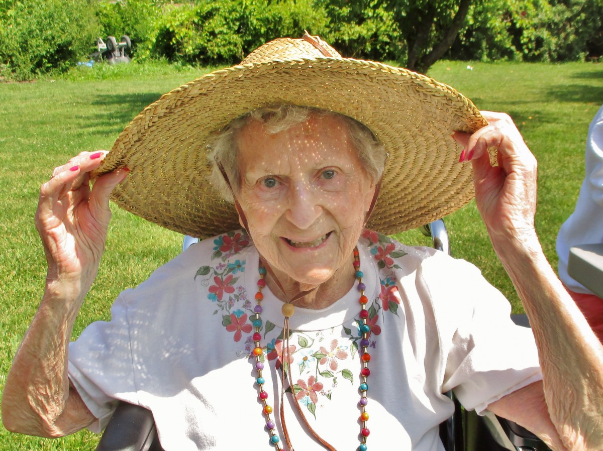 An elderly woman wearing a straw hat and smiling outdoors in a garden on a sunny day.