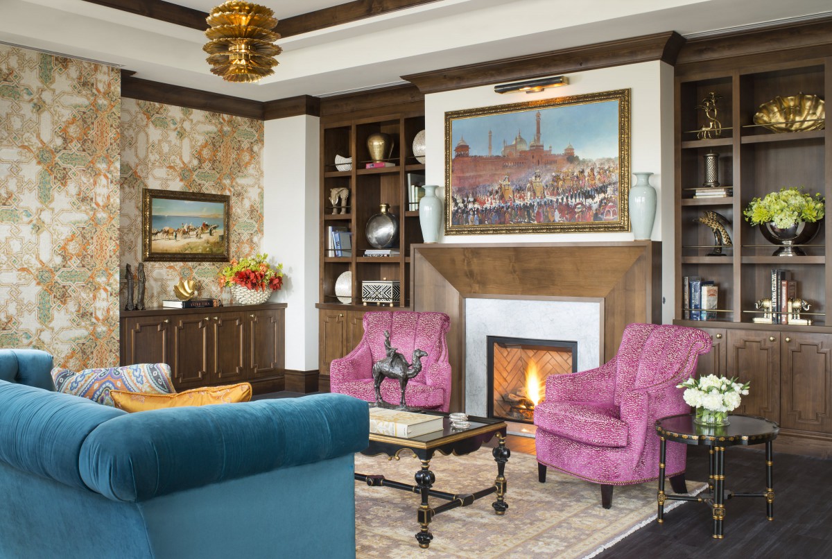 A cozy living room with a fireplace, surrounded by bookshelves and decorated with colorful armchairs and artwork.