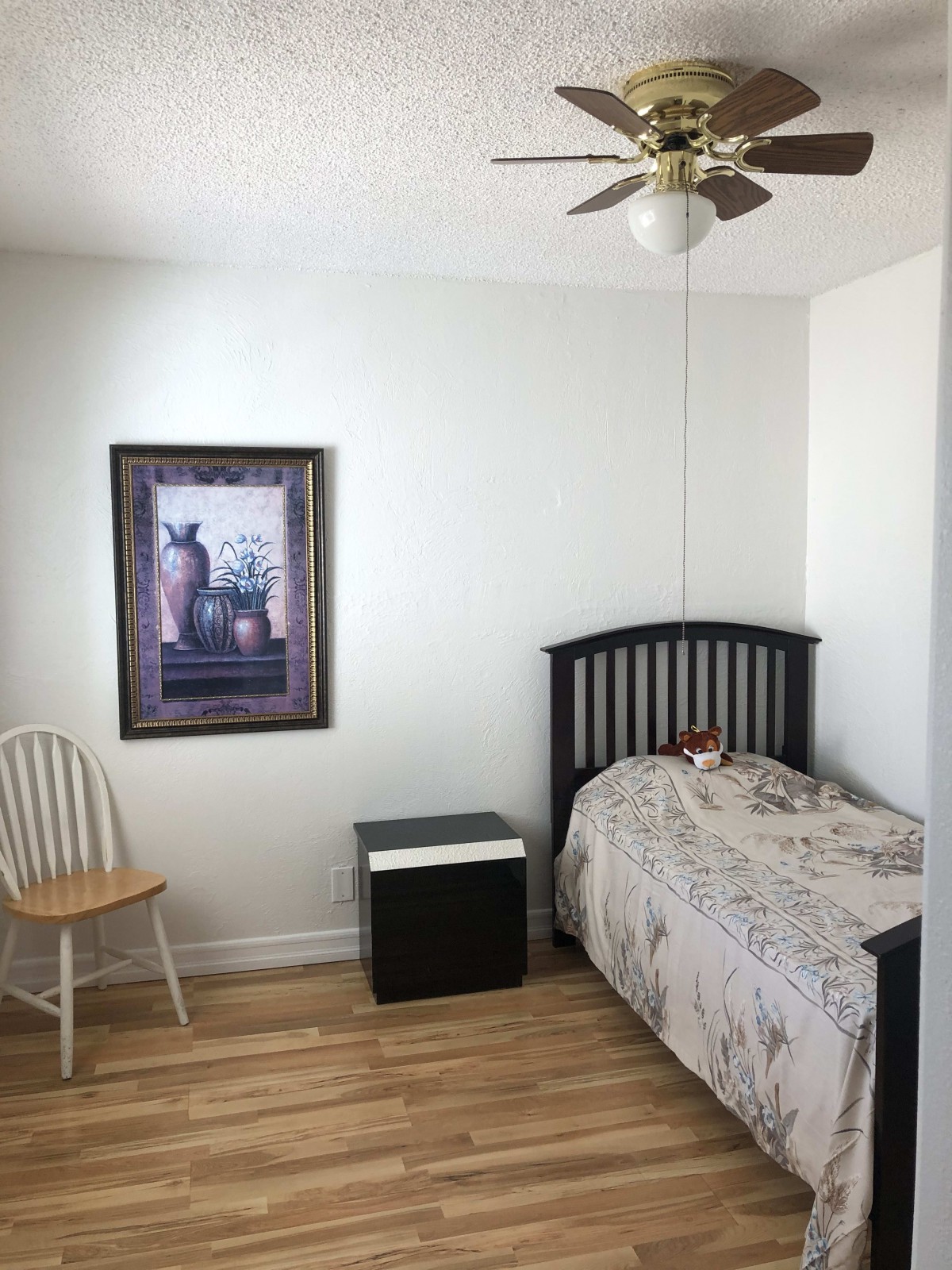 A minimalist bedroom with a single bed, a chair, framed artwork, and a small black side table on a wooden floor.