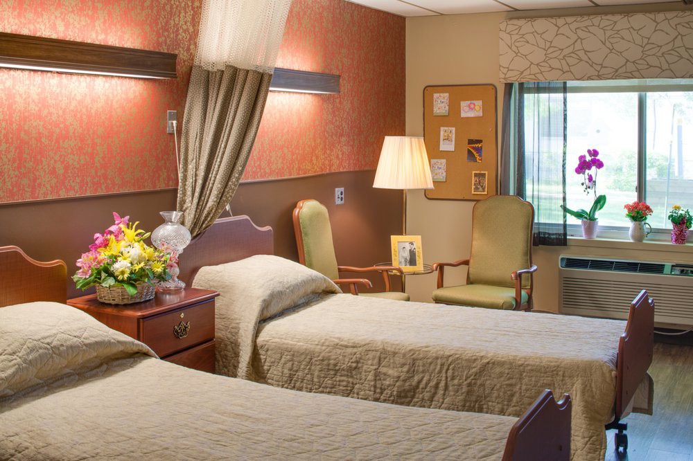 A well-arranged hospital room with two beds, accompanied by bedside furniture, a lamp, flowers, and a window providing natural light.