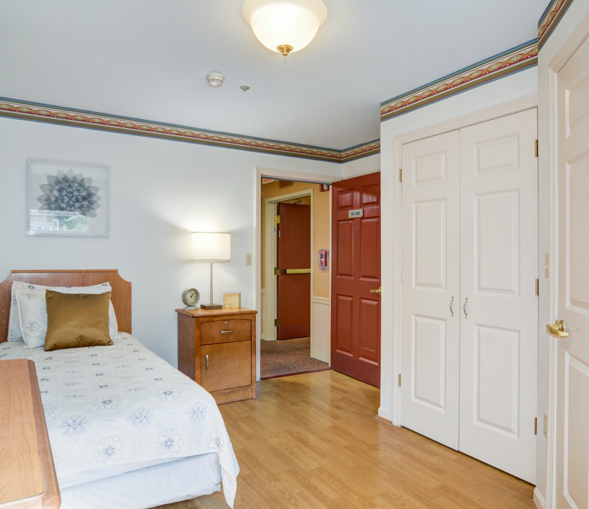 A cozy bedroom featuring a single bed with a nightstand and lamp, wood flooring, and a decorative painting on the wall.