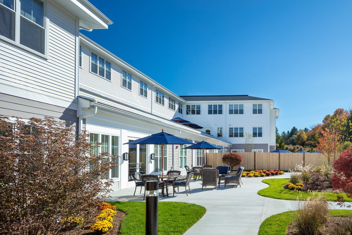 A modern building with a sunlit patio area featuring tables, chairs, and umbrellas.