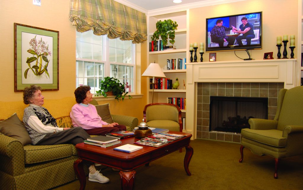 A cozy living room with a person watching television. The room features a fireplace, sofa, coffee table, and bookshelves filled with books and decor.