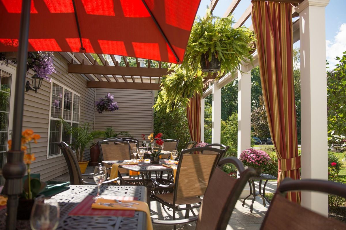 An inviting outdoor patio with tables and chairs under a red umbrella, surrounded by greenery.
