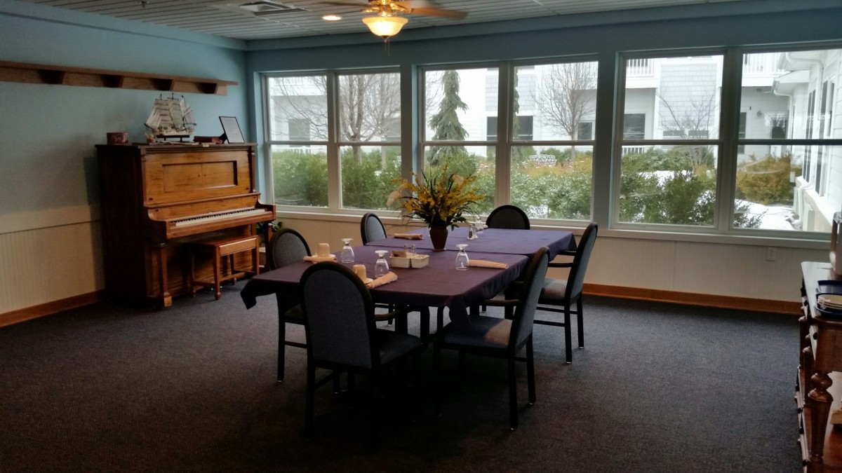 A cozy dining room with a table set for a meal, adjacent to a piano and large windows providing natural light.