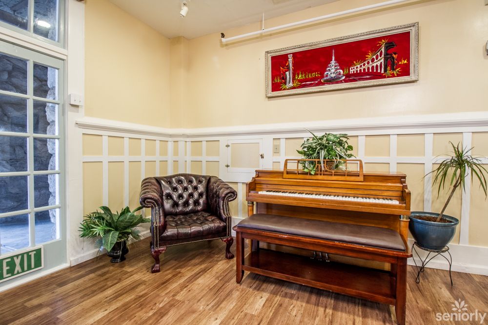 A cozy room with a wooden piano and leather armchair, featuring decorative plants and artwork.