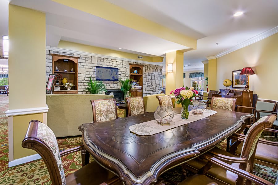 A cozy dining room featuring a large wooden table, upholstered chairs, built-in shelves, and decorative flowers.