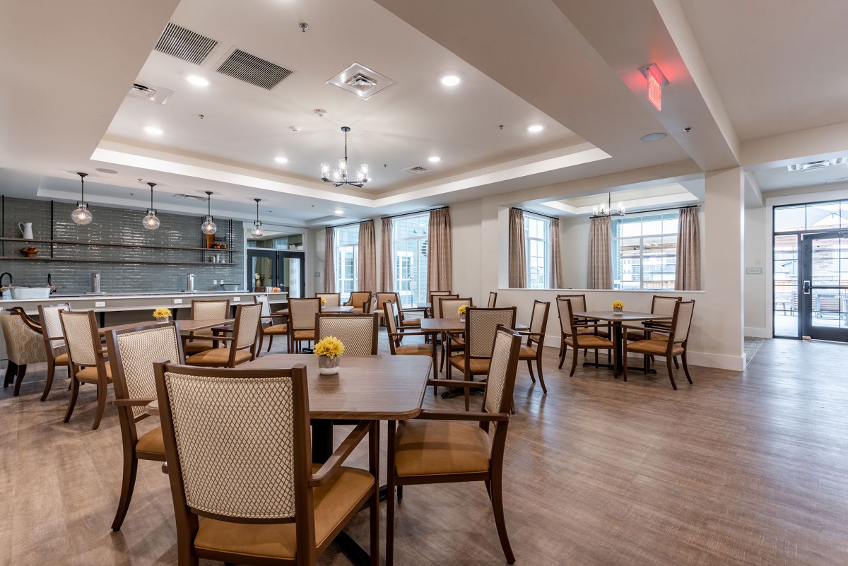 A spacious dining area with wooden flooring, elegantly arranged tables and chairs, and ample natural light from large windows.