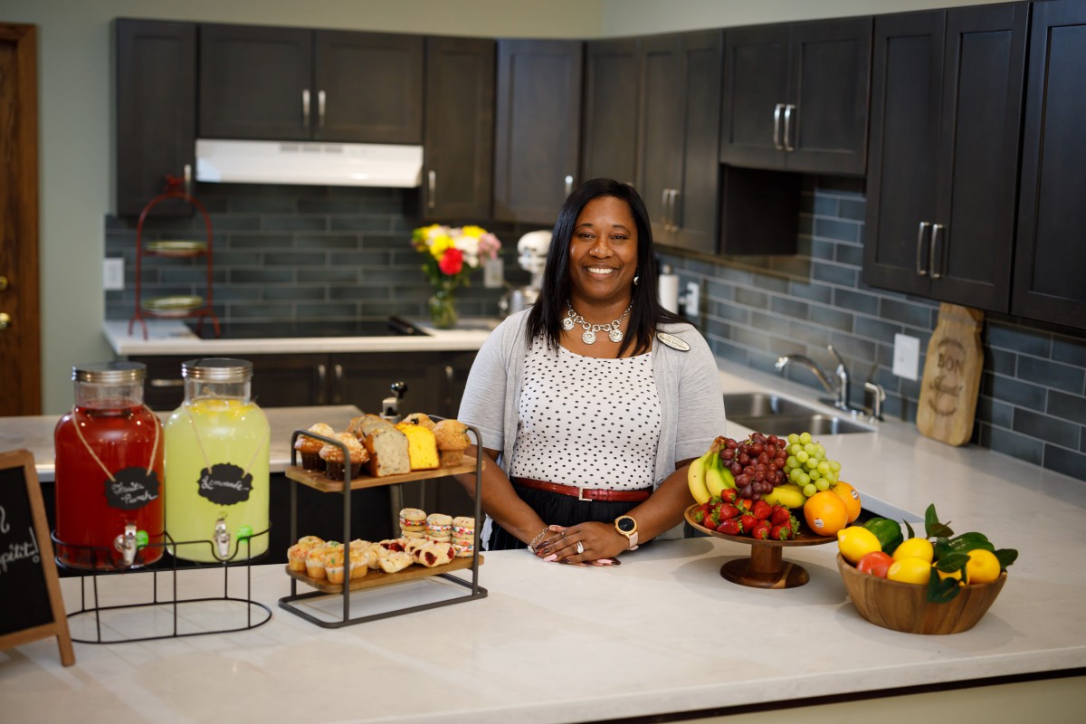 A woman standing in a kitchen surrounded by a display of fruit, baked goods, and lemonade.