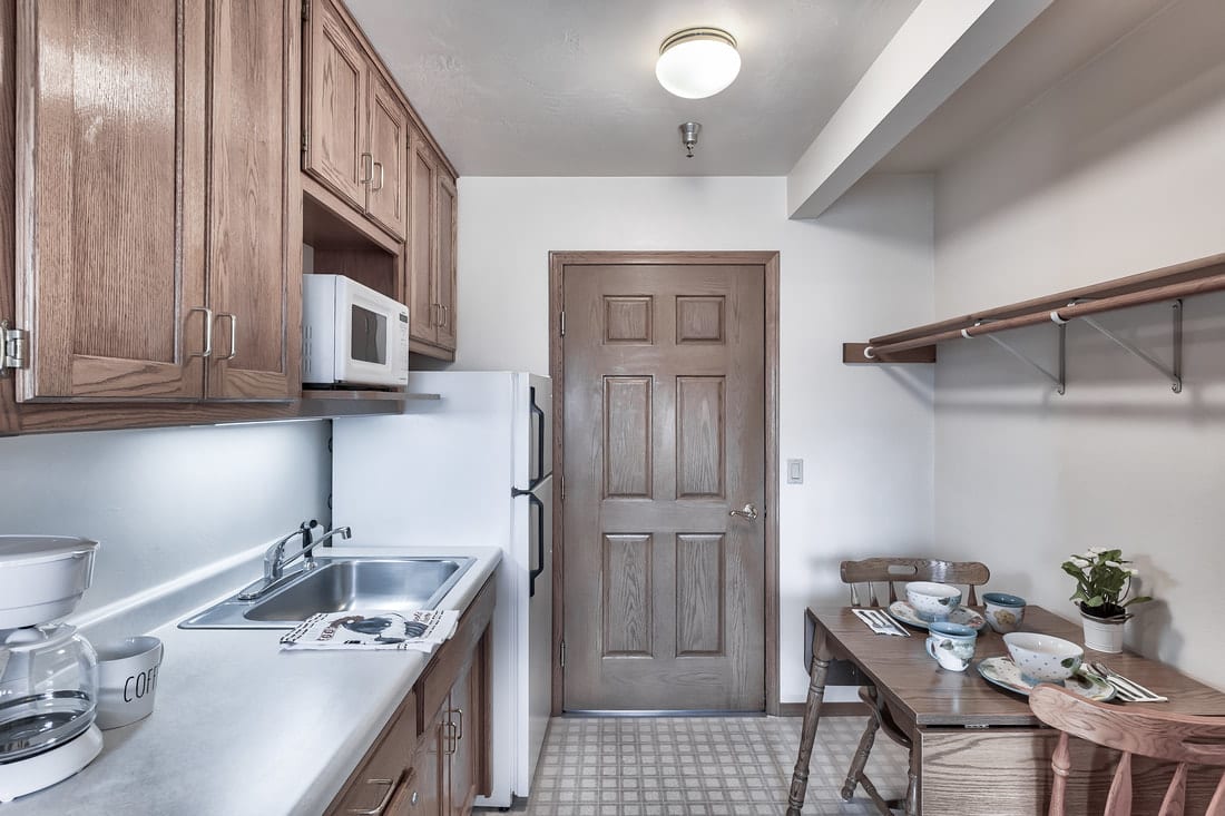 A compact kitchen with wooden cabinets, microwave, refrigerator, sink, and a small dining area.