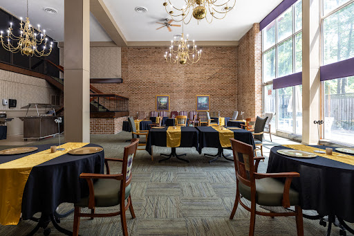 An elegantly arranged dining area featuring tables covered with black cloths and yellow napkins, complemented by a bright ambiance from large windows and decorative chandeliers.