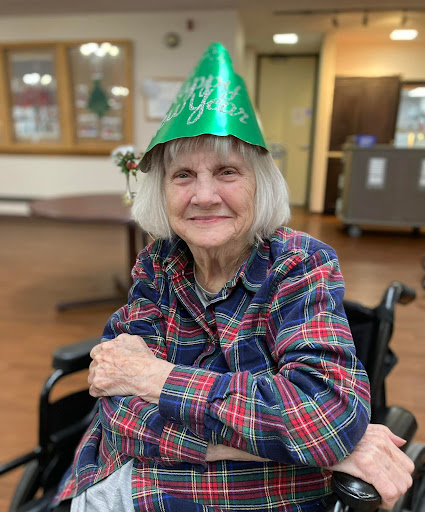 A cheerful elderly woman wearing a green party hat, smiling while seated in her wheelchair.