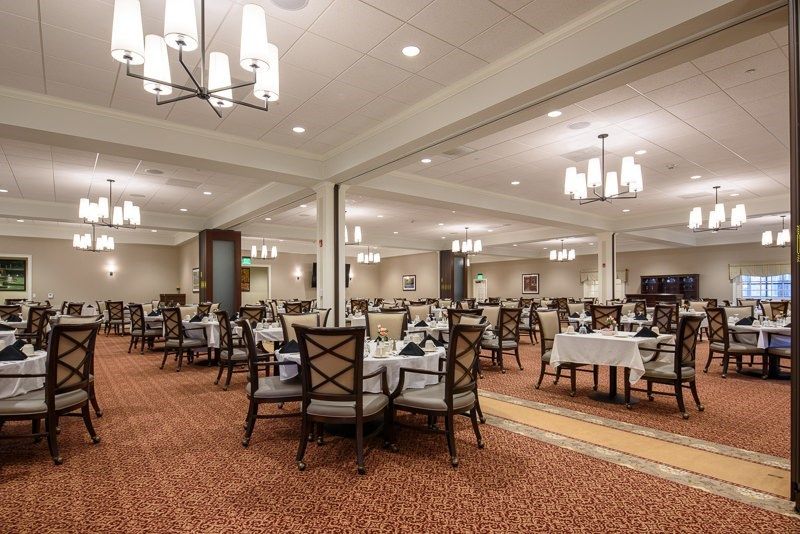 A spacious and elegantly decorated dining room with neatly arranged tables and chairs, featuring warm lighting from chandeliers.