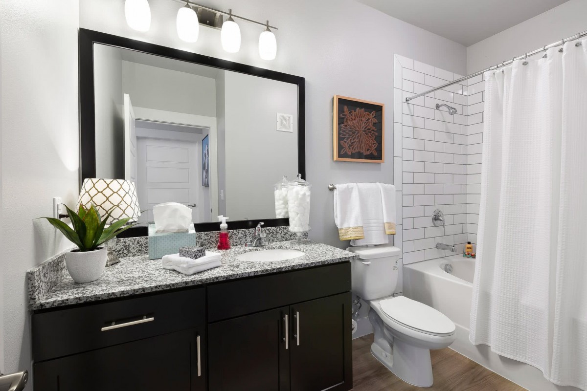 A modern bathroom with dark cabinetry and a marble countertop, featuring a large mirror and a bathtub with white subway tiles.