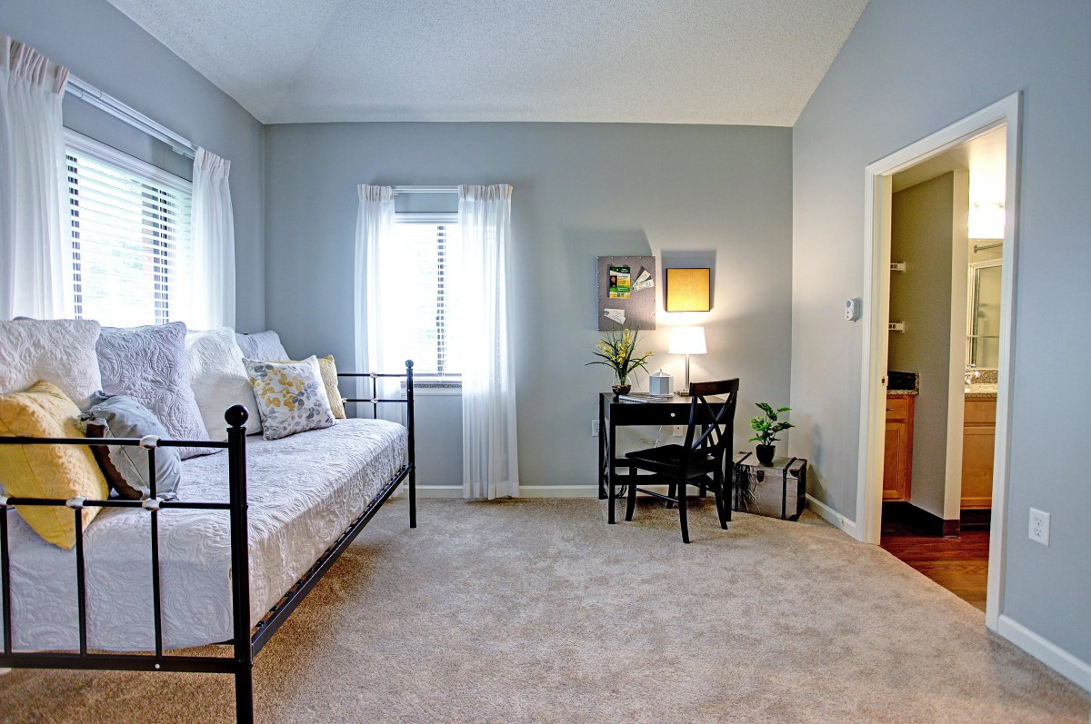 A cozy bedroom featuring a daybed and a small desk, with natural light streaming through the windows.