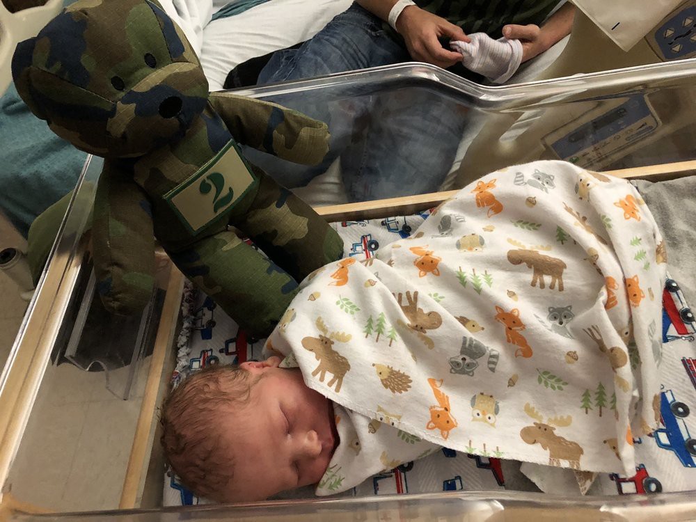 A newborn baby is peacefully sleeping in a hospital bassinet, wrapped in a patterned swaddle blanket next to a teddy bear.