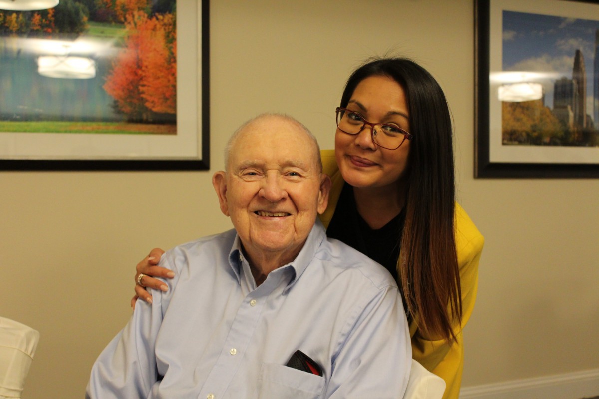 A woman stands behind an elderly man, both smiling warmly.