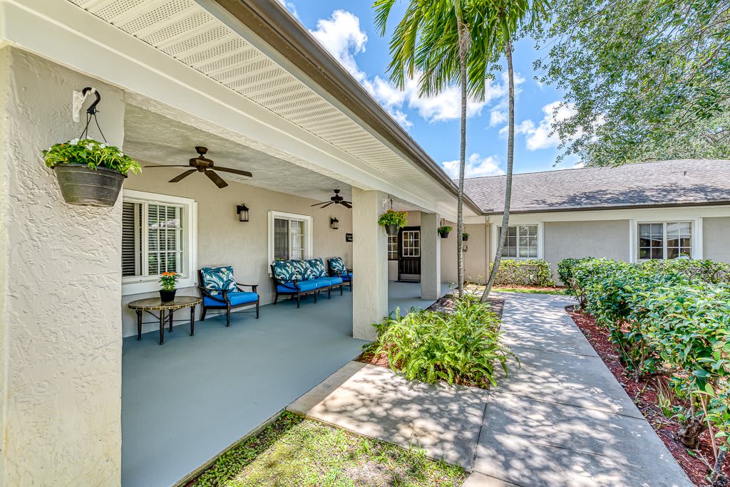 A beautiful house with a covered patio featuring outdoor seating and surrounded by lush landscaping and palm trees.