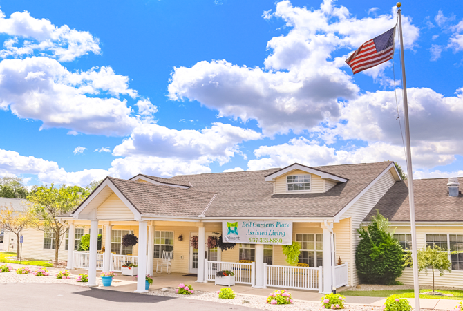 A small house with a sign in front, surrounded by a clear blue sky and clouds, with an American flag waving nearby.