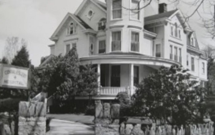 A black and white photograph of a large Victorian-style house with a prominent porch.