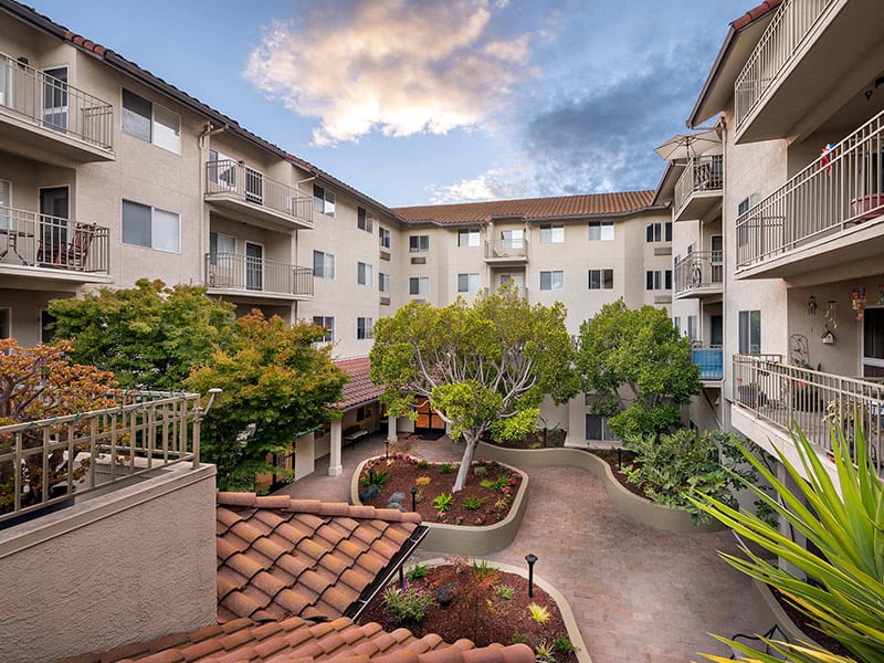 A modern apartment complex with a landscaped courtyard and pathways under a partly cloudy sky.