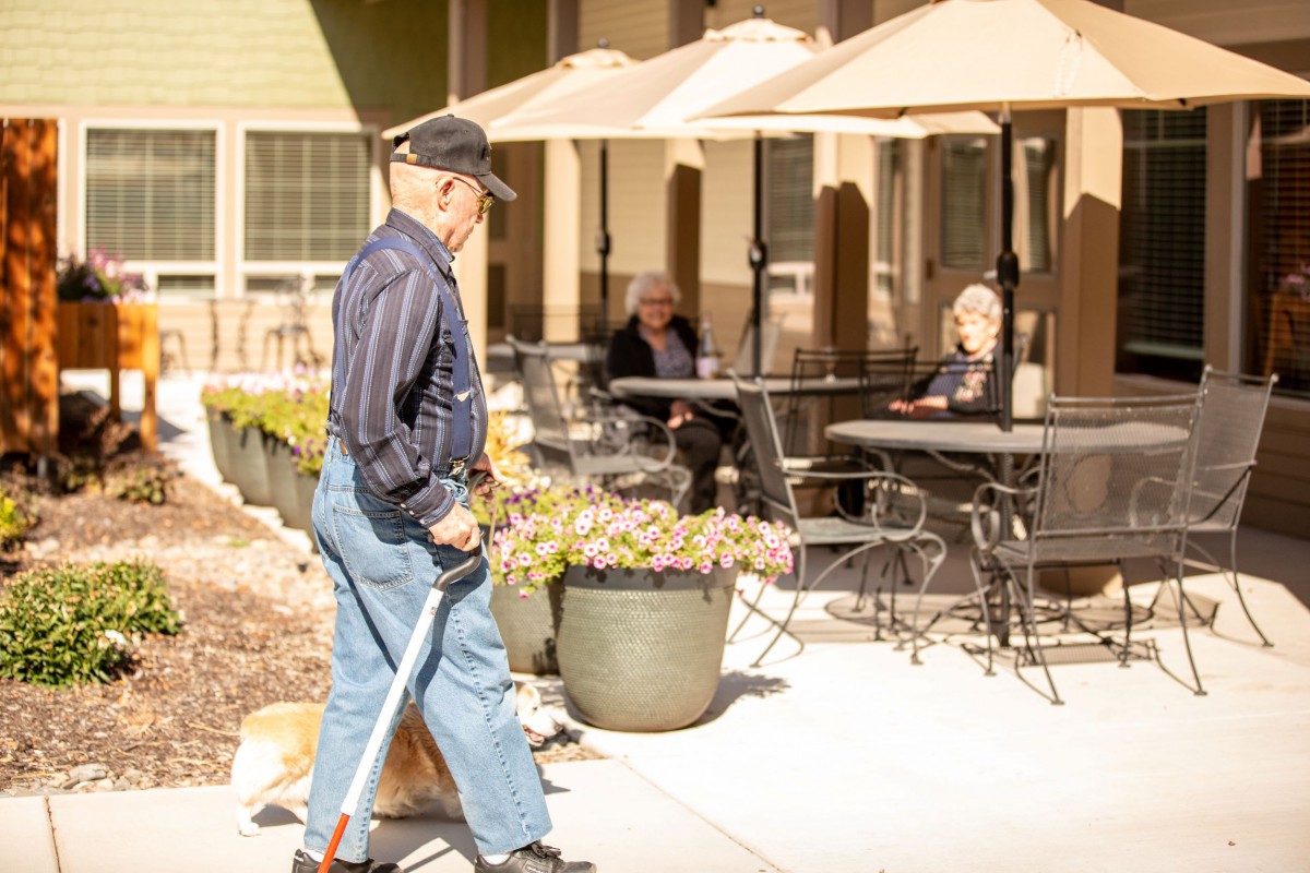 An elderly man walks with a cane past an outdoor patio area under sunny skies.