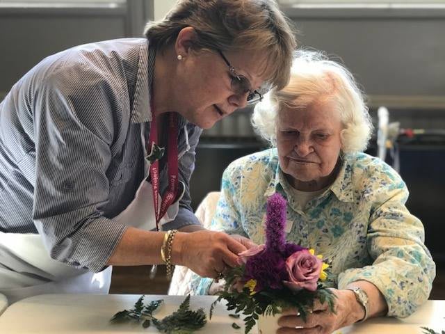 An elderly woman receives assistance in arranging flowers during a craft activity.