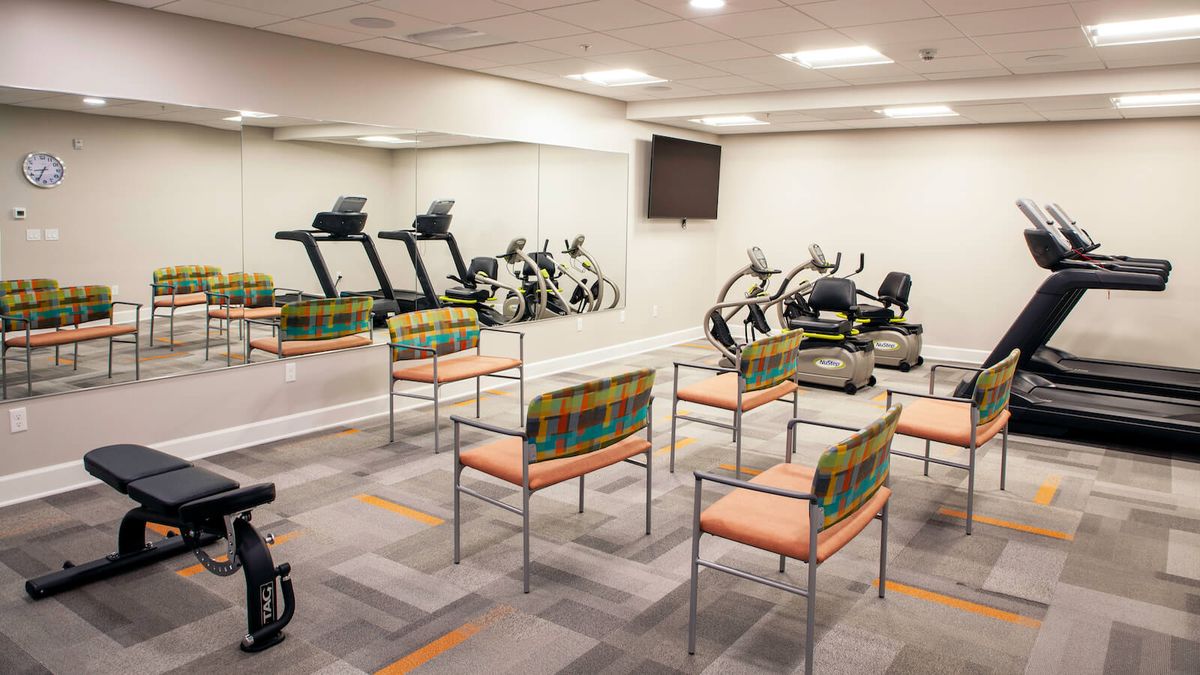 A gym equipped with treadmills, stationary bikes, and chairs, featuring mirrors and a television.