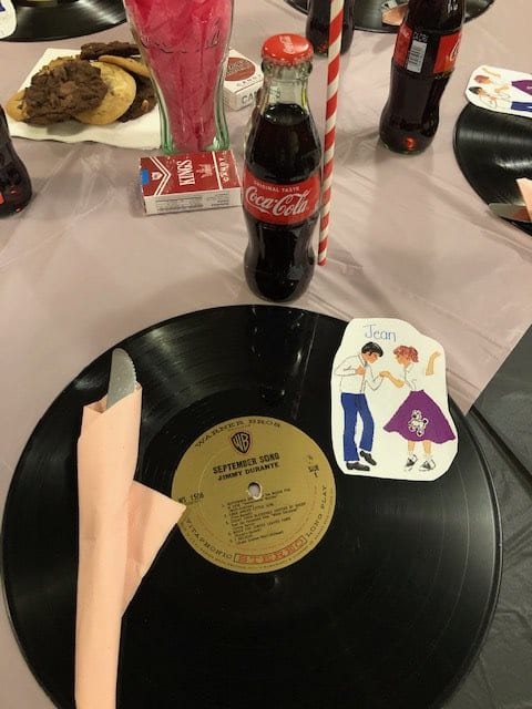 A retro-themed table setting features a vinyl record plate, Coca-Cola bottle with a striped straw, and a vintage dance sticker.