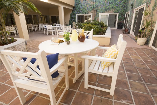 A cozy outdoor patio setup with a white table and chairs adorned with colorful cushions, surrounded by greenery.