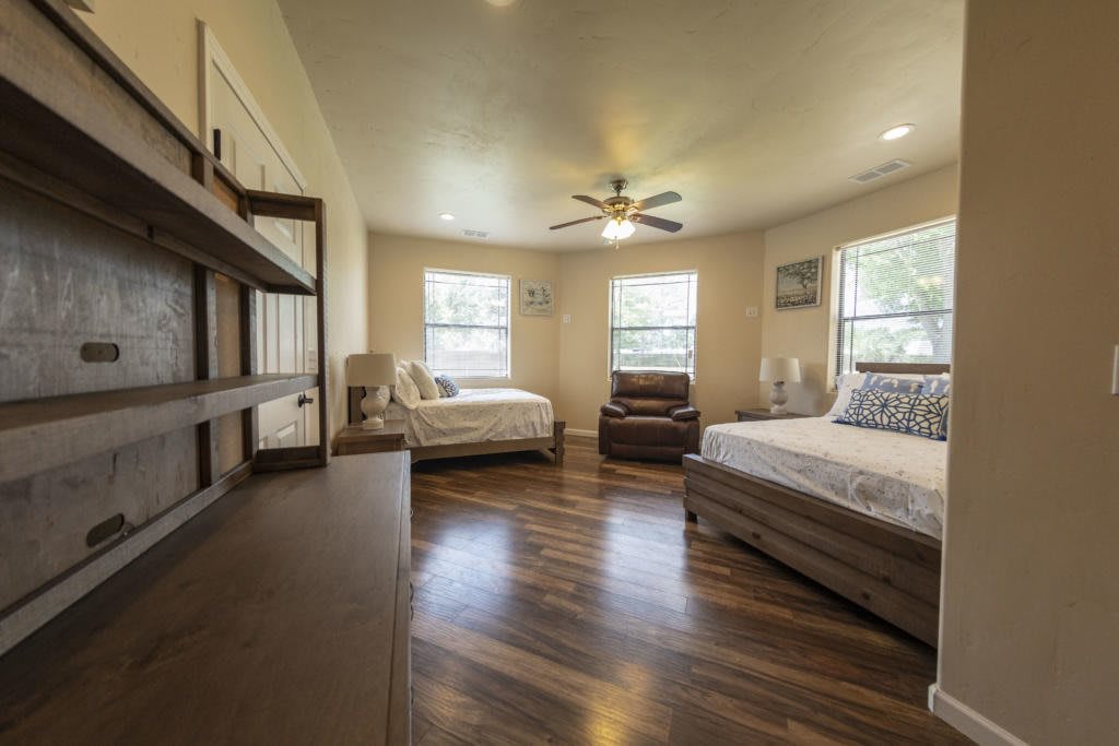 A spacious bedroom with wood flooring, featuring two beds, a ceiling fan, and multiple windows.