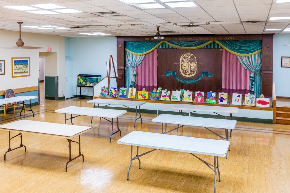 A community center room set up for an art exhibit, featuring paintings displayed on tables in front of a small stage.