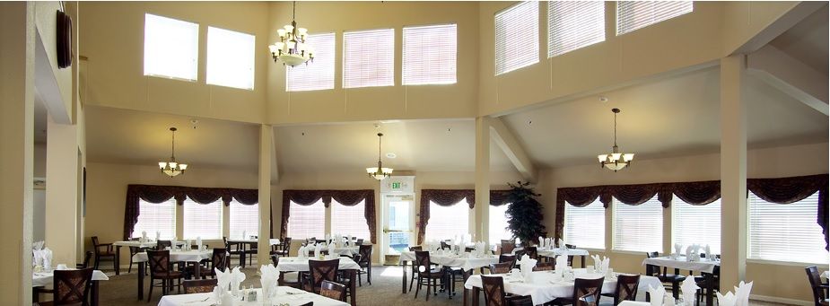 A spacious dining room with elegant chandeliers, neatly arranged tables and chairs, and large windows with curtains.