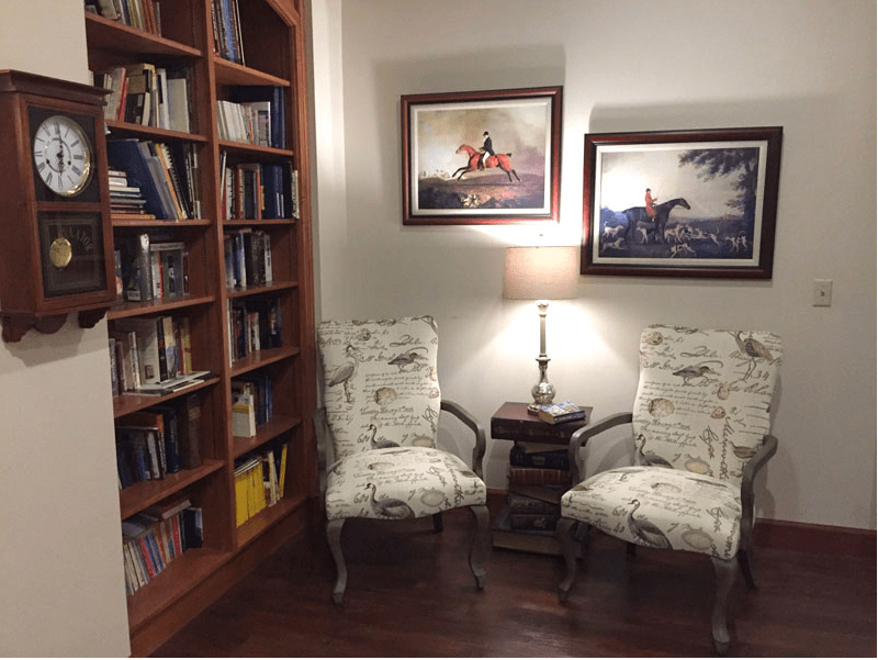 A cozy reading nook featuring two patterned chairs, a wooden side table with a lamp, and surrounded by bookshelves and artwork on the walls.