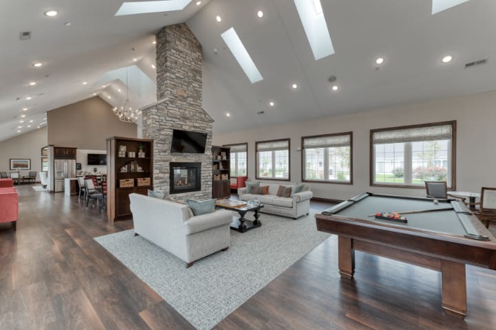 A spacious and modern living room featuring a stone fireplace, comfortable sofas, and a pool table under bright skylights.